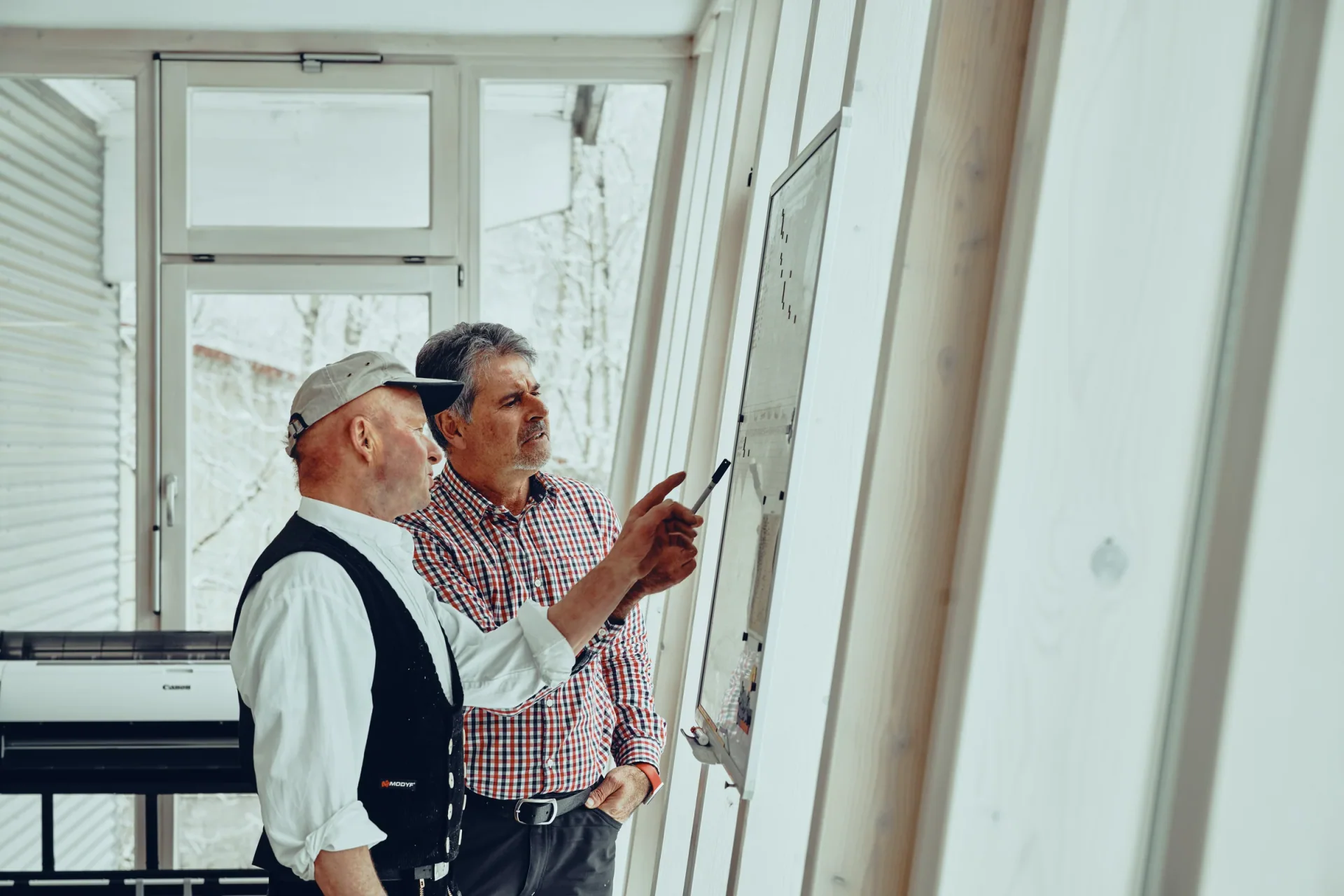 Zwei Zimmerer besprechen einen Plan an der Fensterfront des Büros